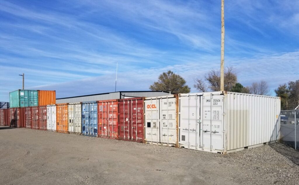 Row of multi-colored shipping containers, including red, orange, white, blue, and green units, lined up along a gravel lot under a blue sky